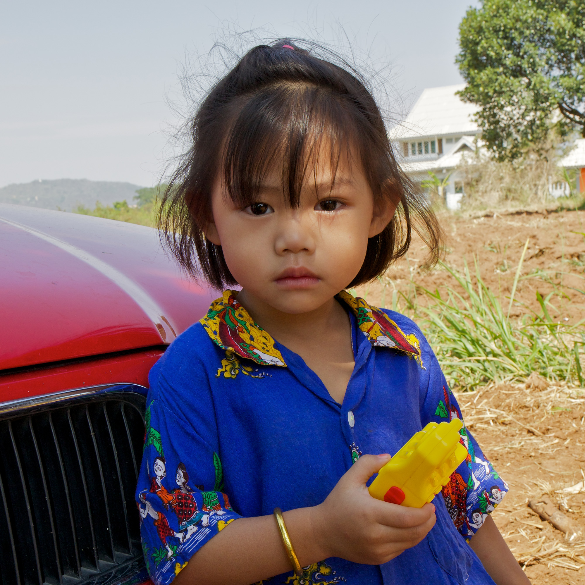 little girl in blue dress, with yellow toy, in front of red car